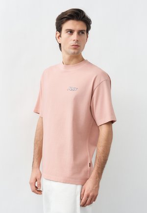 Νέος άνδρας με σκούρα μαλλιά που φοράει ένα ανοιχτό ροζ φαρδύ T-shirt με το λογότυπο «BOSS», στέκεται μπροστά σε ένα απλό ανοιχτό γκρι φόντο.