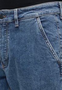 Μπλε τζιν στενή λήψη που δείχνει την μπροστινή τσέπη, ζώνη με θηλιά και μαύρο κουμπί κλεισίματος.
