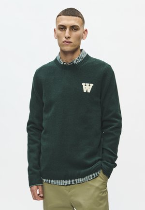 Ung mand med kort hår iført en mørkegrøn sweater med en hvid "W" på brystet over en ternet skjorte og beige bukser.