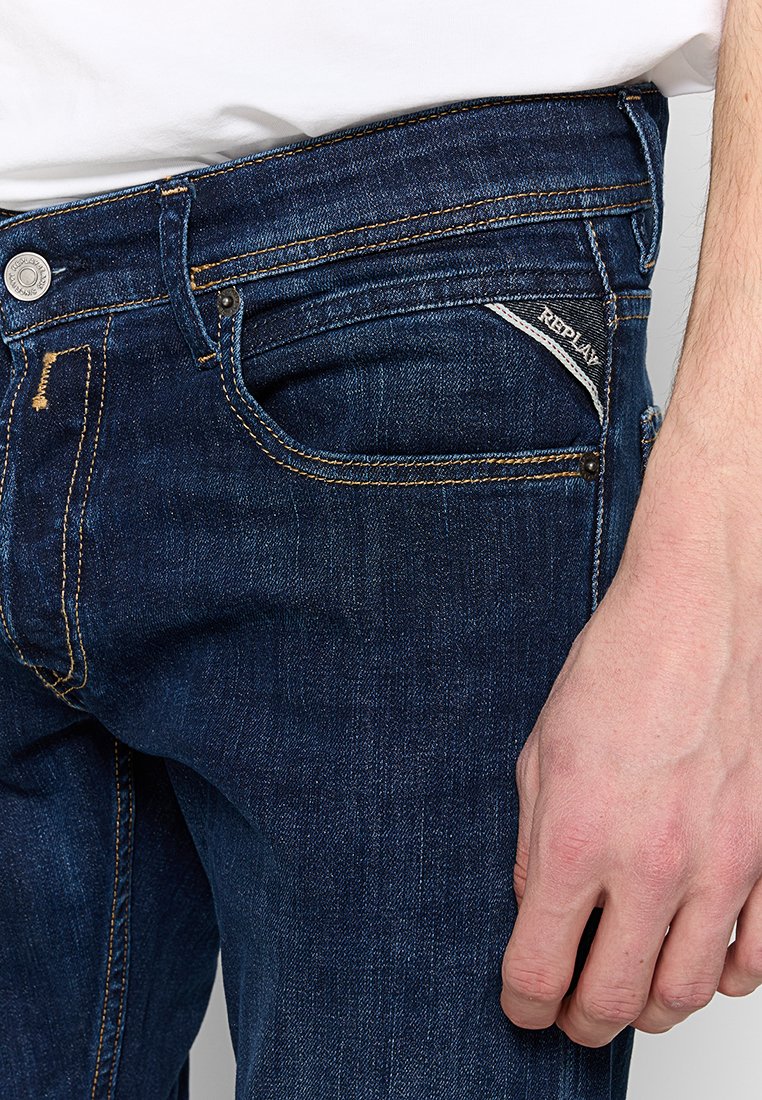 Jeans in denim blu scuro con cuciture arancioni, caratterizzati da un classico design a cinque tasche, un bottone metallico e un logo sulla tasca posteriore.