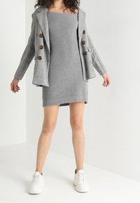 Graues, geripptes Strickkleid mit figurbetontem Schnitt, kombiniert mit einem grauen Karo-Blazer mit runden Knöpfen, vor einem neutralen Hintergrund.