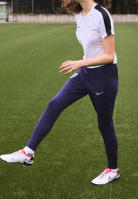 Biała koszulka sportowa z czarnymi akcentami, granatowe spodnie dresowe oraz białe korki z różowymi sznurowadłami, stojące na zielonym boisku.