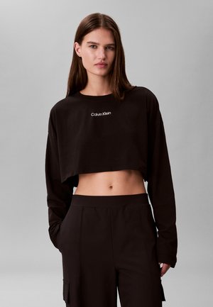 Ung kvinde med glat brunt hår iført sort crop-top med lange ærmer og "Calvin Klein"-logo samt matchende sorte højtsiddende bukser.