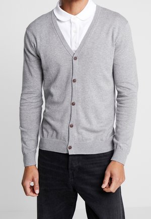 Man draagt een lichtgrijze, geknoopte V-hals cardigan over een wit overhemd met kraag en zwarte broek, staand tegen een effen achtergrond.