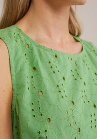 Femeie îmbrăcată cu un top fără mâneci de culoare verde deschis, brodat, cu modele de decupaj circulare, care expune gâtul, umărul și o parte din braț.