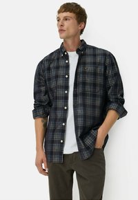 Marineblauwe geruite shirt met witte en grijze accenten, met een knoopsluiting bij de kraag, opgerolde mouwen en een zak op de linkerkant van de borst. Draagt over een witte t-shirt.