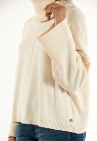 Jersey de cuello alto crema con puños anchos de canalé, combinado con jeans azules, mostrando una mano levantando el cuello cerca de la cara.
