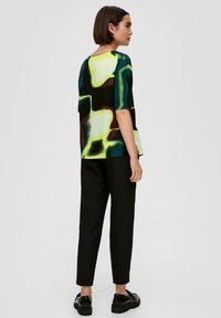 T-shirt à manches courtes avec des motifs abstraits verts, jaunes et bruns, associé à un pantalon noir fuselé et des chaussures noires, vu de dos.