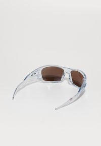 Przezroczyste plastikowe okulary przeciwsłoneczne z brązowymi soczewkami, o eleganckich krzywiznach i charakterystycznym kształcie. Posiadają niebieskie akcenty na oprawie i zausznikach.