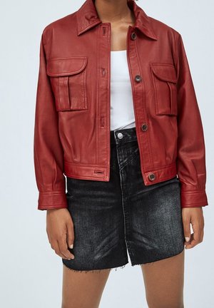 Personne portant une veste en cuir rouge avec poches avant, un haut blanc et une jupe en denim noire avec ourlet effiloché, sur un fond uni.