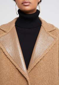 Abrigo texturizado en tono beige con un cuello brillante color crema, superpuesto a un elegante suéter de cuello alto negro. Presenta solapas anchas y un aspecto acogedor.
