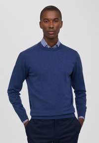 Maglione blu navy a maglia con scollo rotondo e polsini a costine, indossato sopra una camicia a righe blu chiaro e bianco, con una texture liscia.