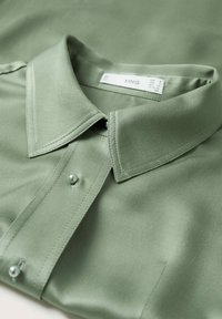Blusa verde satinata con colletto a punta, chiusura frontale con bottoni e bottoni simili a perle. Texture liscia e cuciture rifinite con precisione.