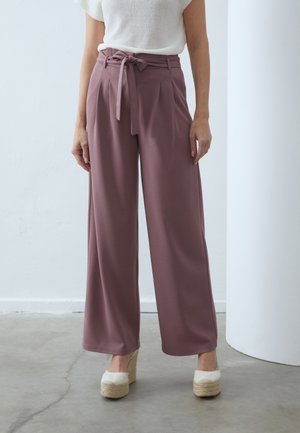 Femme portant un pantalon large taille haute mauve avec une ceinture nouée, un top blanc à manches courtes et des chaussures compensées beiges, debout sur un sol en béton.