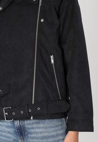 Black faux leather jacket with a belted waist, silver zippers, buttoned collar, and textured finish. Features side zip pocket and lapel accents.