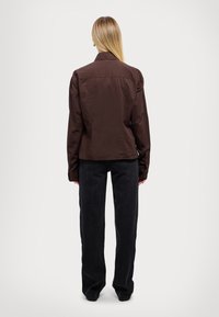 Chaqueta marrón ajustada con cuello, mangas largas y puños con botones, combinada con pantalones negros de corte holgado. Textura suave, diseño minimalista.
