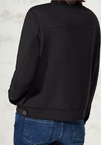 Schwarze Jacke mit einem schlichten Design, versehen mit einem runden Logo-Akzent nahe dem Saum, kombiniert mit blauen Jeans. Sanfte Textur und normaler Schnitt.