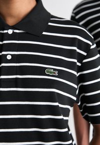Polo din bumbac cu dungi alb-negru, cu guler, având un logo brodat de crocodil verde pe partea stângă. Placket cu trei nasturi.