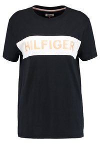 Černé bavlněné tričko s krátkým rukávem, které má na hrudi bílý a oranžový horizontální pruh s nápisem "HILFIGER" vytištěným tučnými písmeny.