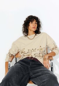 Sweatshirt beige avec broderie florale, associé à un jean gris foncé et une ceinture bordeaux. Les accessoires incluent un bracelet en chaîne dorée chunky et des boucles d'oreilles.