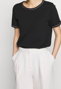 Schwarzes T-Shirt mit kurzen Ärmeln und Rundhalsausschnitt, mit weißen Pipings entlang des Ausschnitts und der Ärmel, kombiniert mit hellen Hosen.