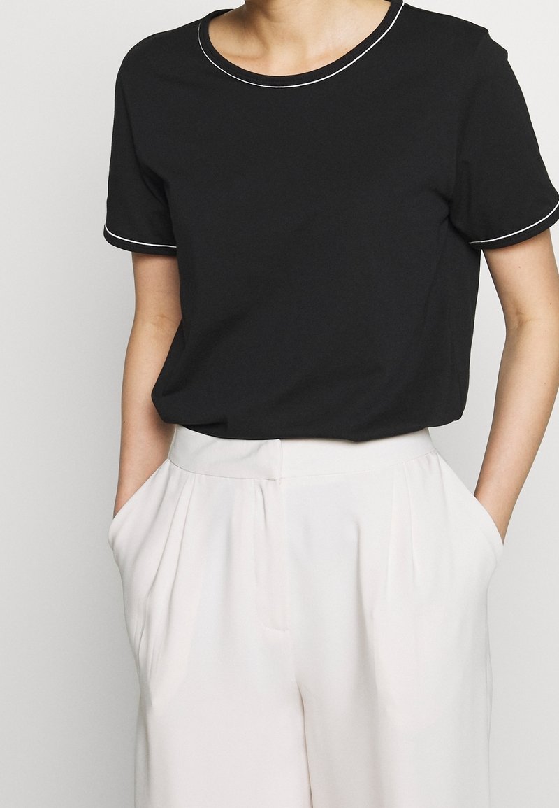 Schwarzes T-Shirt mit kurzen Ärmeln und Rundhalsausschnitt, mit weißen Pipings entlang des Ausschnitts und der Ärmel, kombiniert mit hellen Hosen.