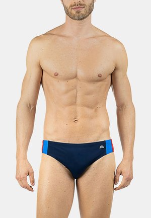 Uomo a torso nudo che indossa slip da bagno blu navy con pannelli laterali rossi e blu, in piedi contro uno sfondo bianco semplice.