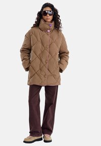 Brązowa pikowana kurtka z wysokim kołnierzem, różowymi akcentami i dużymi guzikami. W połączeniu z brązowymi spodniami i jasnymi butami.