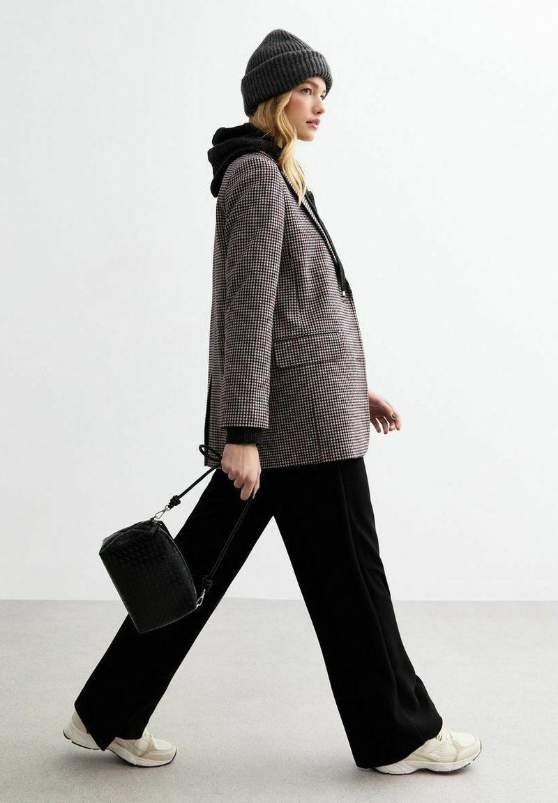 Blazer a quadri grigio e nero, indossato sopra un hoodie nero, abbinato a pantaloni neri a gamba larga; accessoriato con una piccola borsa nera e un berretto grigio.