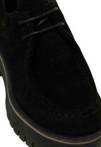 Zapato de ante negro con punta redonda, detalles de cordones y una suela de goma gruesa que presenta una textura estriada y un acento marrón en contraste.