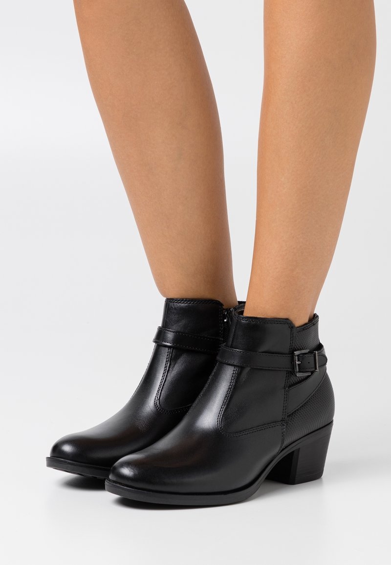 Bottines en cuir noir avec des sangles à boucle sur le côté et des talons blocs portées sur des jambes nues sur un fond uni.