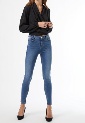 Vrouw die een zwarte lange mouwen wikkelblouse draagt, blauwe skinny jeans en zwarte puntige hakken, staand tegen een witte achtergrond.