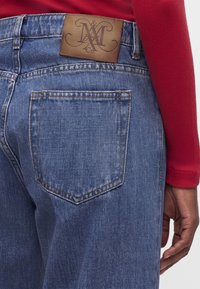 Οπίσθια όψη μπλε τζιν από denim με δερμάτινο λογότυπο πάνω από την δεξιά πίσω τσέπη, φορεμένα με κόκκινο μακρυμάνικο μπλουζάκι και εμφανές χέρι.