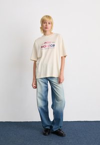 T-shirt surdimensionné crème avec le logo "new balance" en rouge et bleu, associé à un jean bleu délavé à jambes larges et des chaussures noires à lacets.
