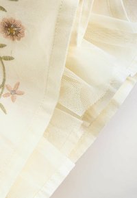 Tissu couleur crème avec broderies florales, doté de couches en mesh délicates et d'une texture lisse, mettant en valeur des points de couture complexes et de douces fronces.