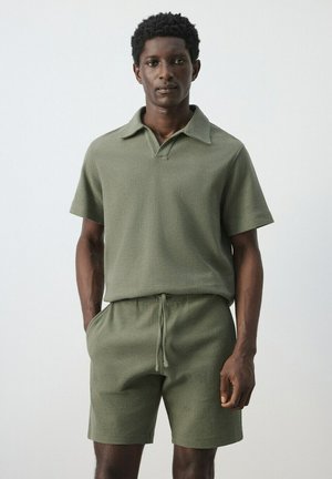 Hombre de pie con las manos en los bolsillos, vistiendo una camisa polo de punto color verde oliva de manga corta y unos pantalones cortos de punto a juego, contra un fondo claro y liso.