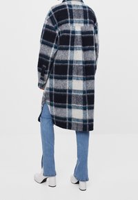 Kostkovaný kabát z vlněné směsi v námořnické, světle šedé a tyrkysové barvě, s volným střihem, dlouhými rukávy a bočními rozparky, nošený přes rozšířené džíny a lodičky.
