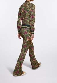 Chaqueta con cremallera de flores y pantalones acampanados en tela verde con flores moradas y naranjas, con puños y cintura acanalados, combinados con zapatillas blancas.