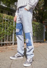 Vaqueros de mezclilla de color azul claro con acentos de parches en azul oscuro y detalles desgastados, combinados con zapatillas de deporte en blanco y negro.