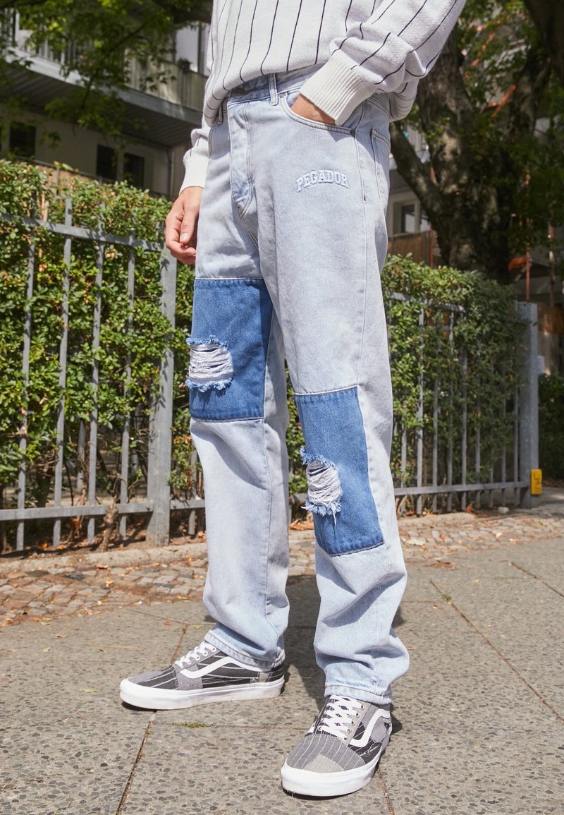 Vaqueros de mezclilla de color azul claro con acentos de parches en azul oscuro y detalles desgastados, combinados con zapatillas de deporte en blanco y negro.