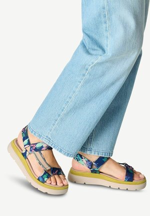 Füße in hellblauen Jeans und bunten blauen Sandalen mit Blumenmuster und dicken beigen Sohlen auf hellem Hintergrund.