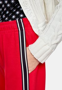 Red jogger pants with black and white side stripes, paired with a white cable-knit sweater. Fabric: soft, elastic waistband.