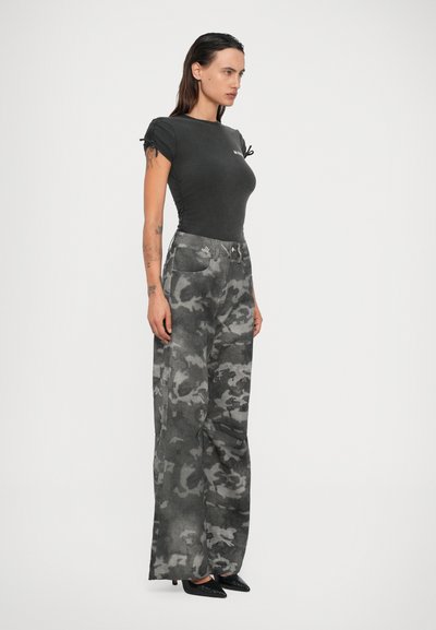 Femme avec de longs cheveux bruns portant un haut noir ajusté, un pantalon large gris camouflage et des escarpins noirs à bout pointu, se tenant de profil.