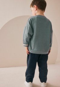 Dječji outfit sastoji se od svijetloplavog duksera s rebrastim detaljima i tamnoplavih trenirki. Tkanina izgleda mekano i ležerno. Bijele tenisice upotpunjuju izgled.