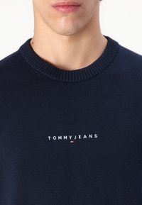 Mornarsko plava pletiva pulover s rebrastim okruglim izrezom. Ima bijeli izvezeni logo "TOMMYJEANS" preko prsnog dijela. Jednostavan dizajn, ležeran kroj.