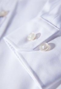 Puño de camisa de algodón blanca con dos botones redondos y transparentes. La tela muestra una textura suave con un pliegue limpio en el borde.