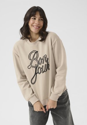 Sweat-shirt beige avec texte brodé "Bon Jour" en noir. Il possède un col rond, des manches longues et des poignets côtelés. Texture de tissu douce.