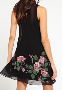 Svart ärmlös klänning med en genomskinlig baksida, prydd med färgglad blommig broderi i rosa och grönt längs fållen, vilket skapar en texturerad design.