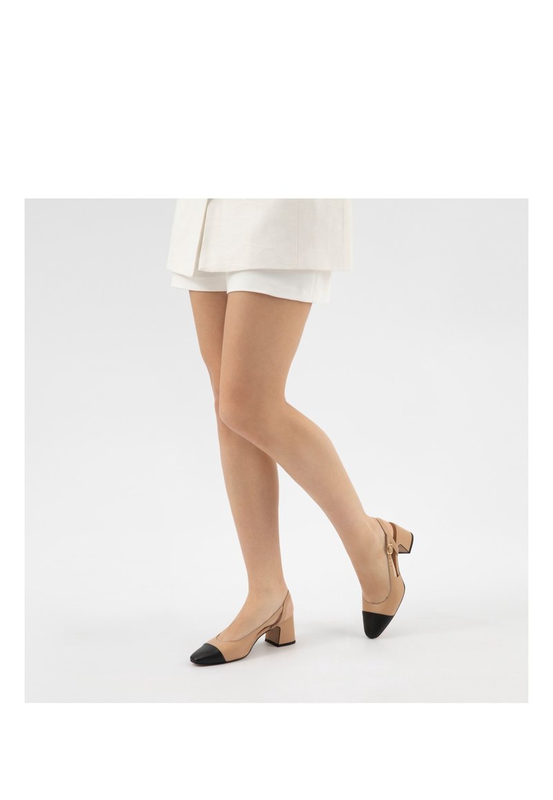 Femme portant des chaussures slingback à talon bloc beige et noir avec une bride à la cheville, associées à un short blanc et un blazer clair sur fond blanc.