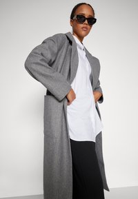 Γκρι μάλλινο παλτό με χαλαρή εφαρμογή, μακριά μανίκια και δύο μπροστινές τσέπες, συνδυασμένο με λευκό πουκάμισο με κουμπιά και μαύρο παντελόνι.
